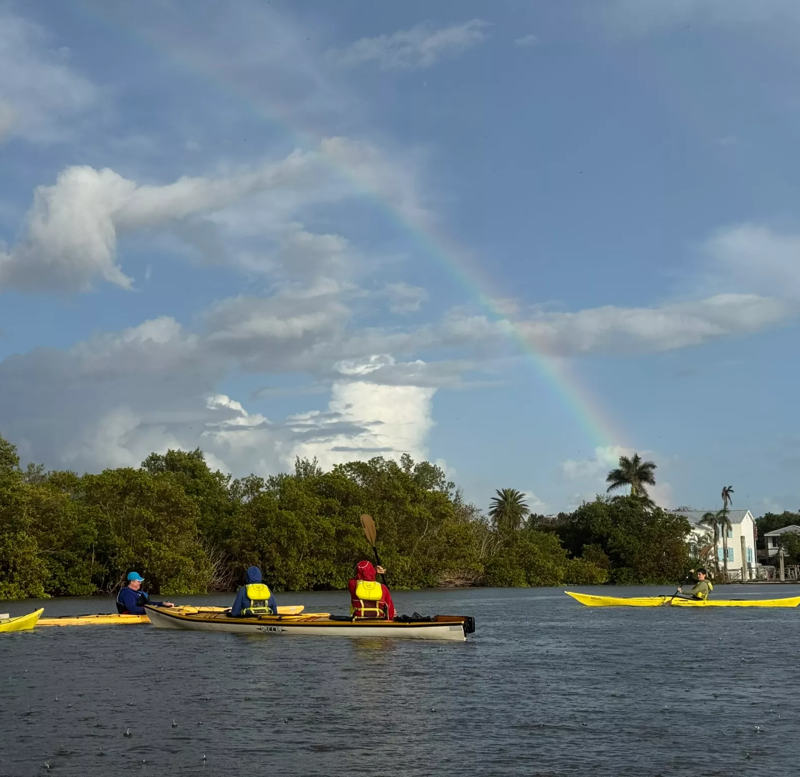 The image shows a group of people kayaking on a body of water, likely a river or bay, under a vibrant rainbow. The kayakers are in yellow and white kayaks, paddling near a lush green shoreline with trees and some houses visible in the background. The sky is filled with dramatic clouds, adding to the scenic beauty of the moment.
